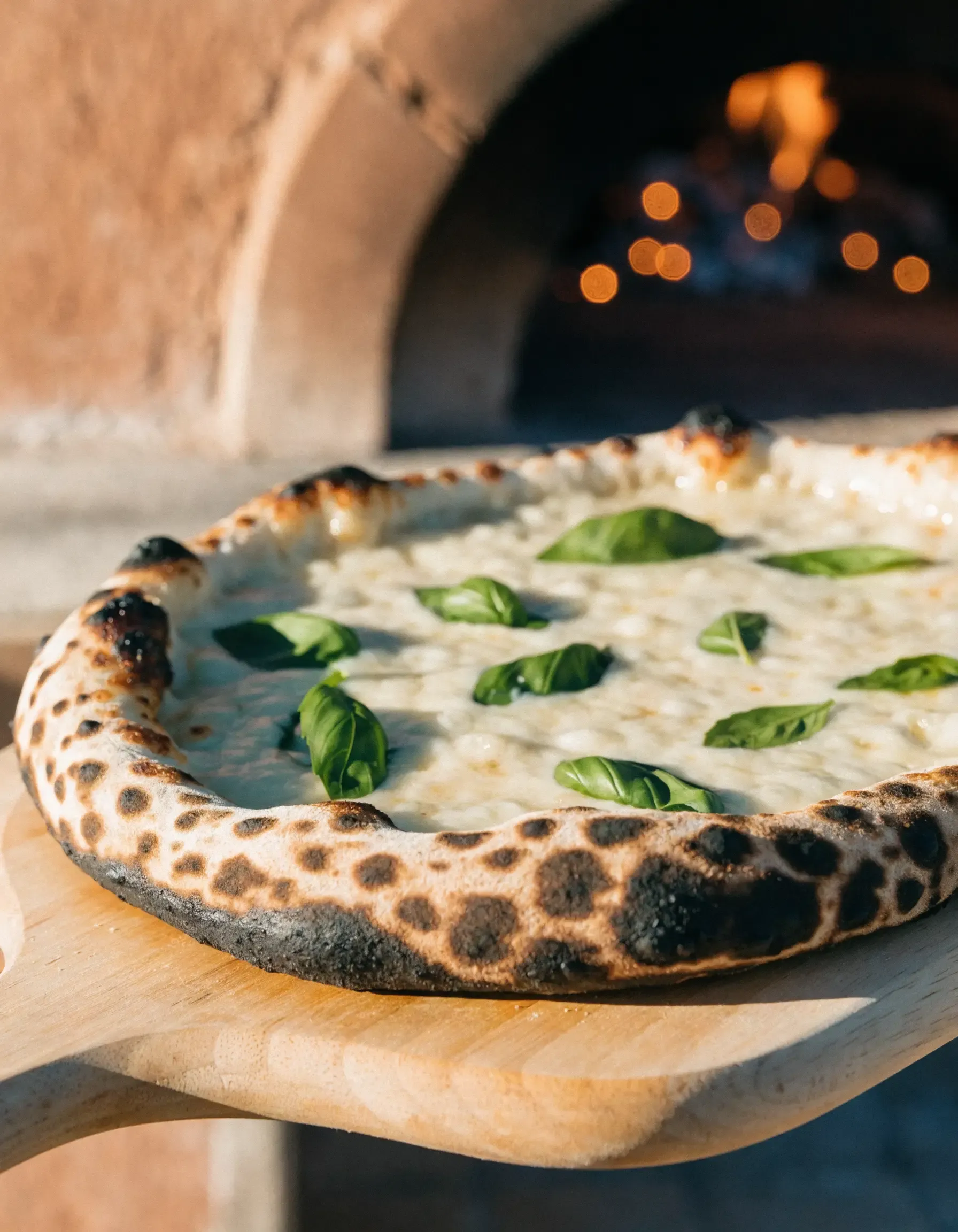 Una pizza appena sfornata sulla pala di legno, davanti alla bocca del forno con il fuoco acceso in fondo